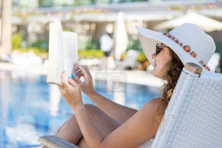 Téléchargez les photos : Jeune femme lecture livre dans chaise longue près de la piscine - en image libre de droit