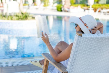 Photo for Young woman reading book in deck chair near swimming pool - Royalty Free Image