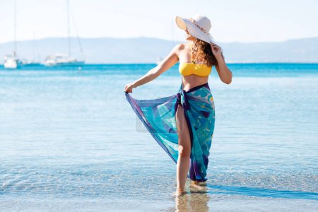 Téléchargez les photos : Jeune femme avec un chapeau de paille dans un maillot de bain jaunâtre et un sarong bleu profite de la plage - en image libre de droit