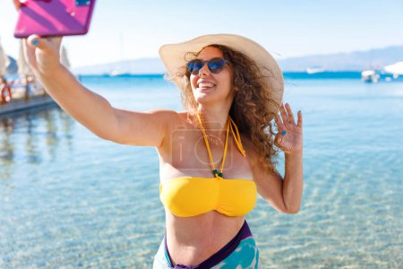 Foto de Mujer joven sonriente con sombrero de paja y gafas de sol tomar una selfie en la playa - Imagen libre de derechos
