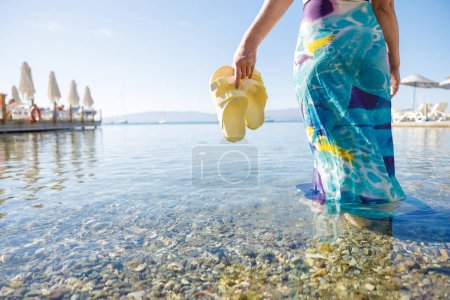Téléchargez les photos : Femme pieds nus tenir sandales jaunes et marcher sur l'été le long de l'eau de mer et le sable sur la plage - en image libre de droit