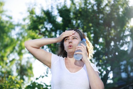 Photo for Young woman having hot flash and sweating in a warm summer day. Woman touching face with cold water bottle to cool off - Royalty Free Image