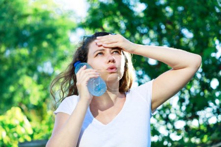 Mujer joven con sofocos y sudoración en un cálido día de verano. Mujer tocando la cara con botella de agua fría para refrescarse