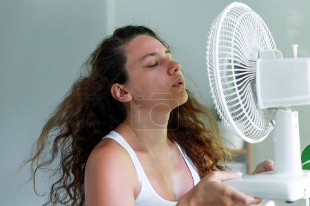 Foto de Mujer joven dentro, tratando de refrescarse delante de un ventilador eléctrico. Ella está de pie junto a una ventana con su cara cerca del ventilador eléctrico - Imagen libre de derechos