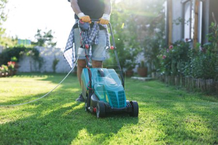 Téléchargez les photos : Jeune homme dans son jardin tondre l'herbe avec une tondeuse à gazon par une journée ensoleillée - en image libre de droit