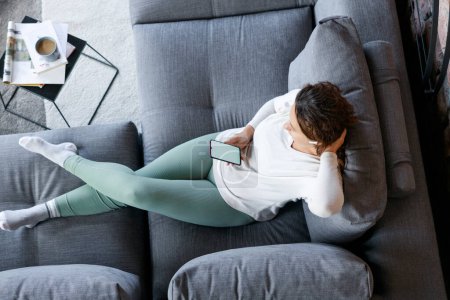 Foto de Mujer joven acostada en un sofá en la sala de estar organizando la lista de reproducción y escuchando música desde la aplicación en su teléfono inteligente - Imagen libre de derechos
