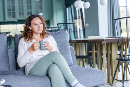 Téléchargez les photos : La jeune femme s'assied sur le canapé en buvant du café. Loisirs, mode de vie pour les personnes concept - en image libre de droit