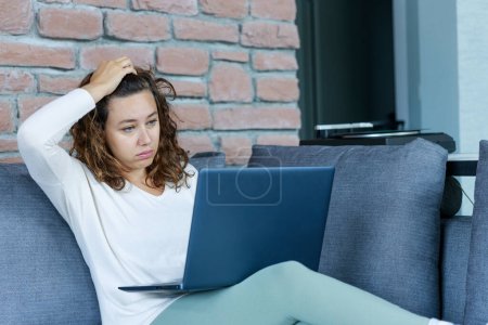 Photo for Young woman with laptop pc sitting at the living room, touching her forehead. Upset woman sitting on sofa, feels unhappy and shocked - Royalty Free Image