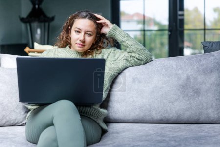 Téléchargez les photos : Jeune femme assise sur le canapé et à l'aide d'ordinateur portable à la maison - en image libre de droit