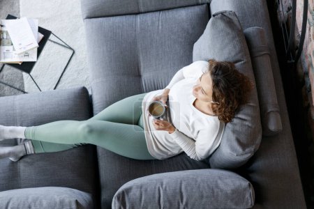 Téléchargez les photos : Jeune femme couchée sur le canapé dans le salon à boire du café et écouter de la musique de écouteurs intra-auriculaires - en image libre de droit