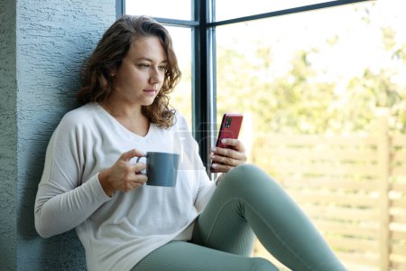 Photo for Young woman holding coffee cup and using phone while sitting at windowsill at home - Royalty Free Image