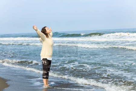 Photo for A woman raising hands while strolling on the beach with the sea and blue sky - Royalty Free Image