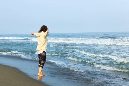 Téléchargez les photos : Une femme levant la main tout en se promenant sur la plage avec la mer et le ciel bleu - en image libre de droit