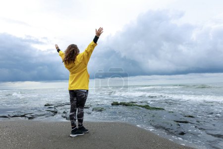 Téléchargez les photos : Jeune femme en imperméable jaune levant les mains contre la mer par un temps orageux et se sentant libre - en image libre de droit