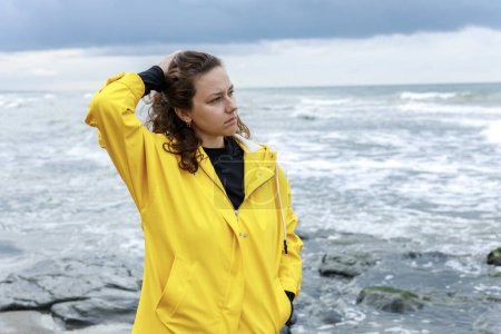 Foto de Mujer joven y nerviosa con impermeable amarillo parada sola junto al mar en un día lluvioso - Imagen libre de derechos