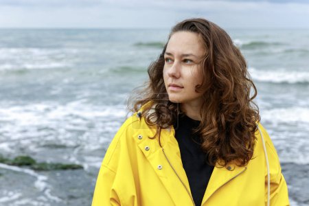 Téléchargez les photos : Triste jeune femme en imperméable jaune marchant seule sur la plage par temps froid nuageux - en image libre de droit