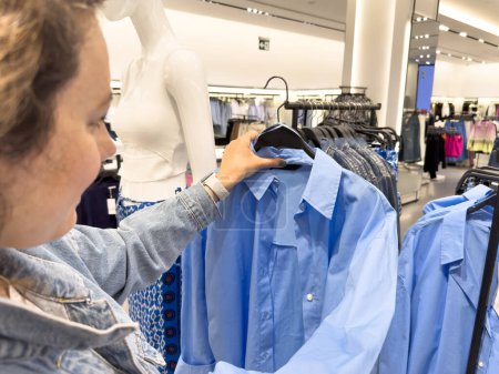 Téléchargez les photos : Jeune femme choisissant des vêtements et regardant chemise bleue sur cintre dans ses mains dans le centre commercial ou magasin de vêtements - en image libre de droit