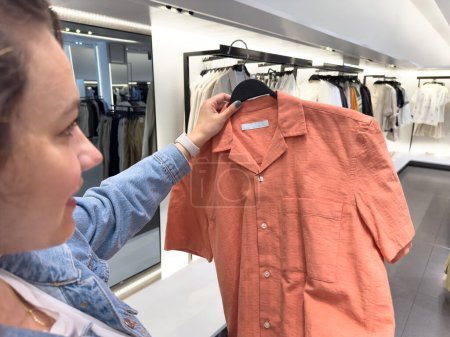 Foto de Mujer joven eligiendo ropa y mirando la camisa en la percha en sus manos en el centro comercial o tienda de ropa - Imagen libre de derechos