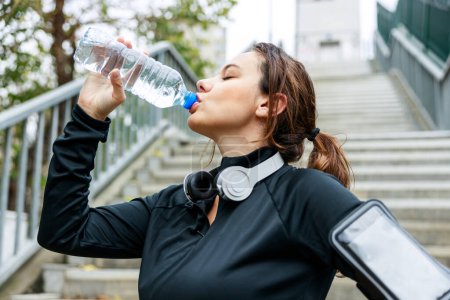 Téléchargez les photos : Jeune sportive buvant de l'eau d'une bouteille après l'entraînement sur fond de ville - en image libre de droit