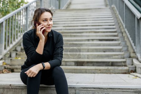 Foto de Una joven sentada en las escaleras y hablando por teléfono. Mujer atleta sonriente tomando un descanso durante el ejercicio en la ciudad. Concepto de deporte, fitness y estilo de vida saludable - Imagen libre de derechos