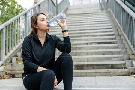 Téléchargez les photos : Jeune femme prenant pause jogging et boire de l'eau d'une bouteille en plein air - en image libre de droit