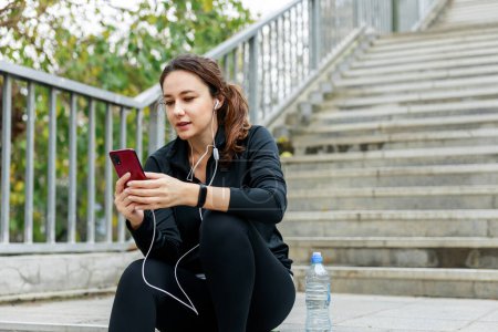 Photo for Young woman taking a break after exercising and listening to music on staircase. Casual active people concept - Royalty Free Image