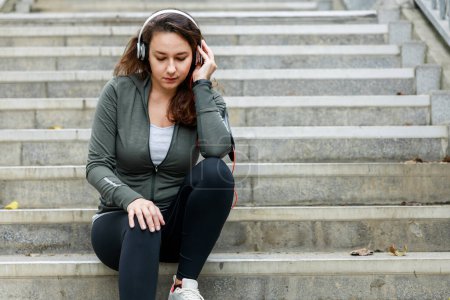 Téléchargez les photos : Jeune femme sportive écoutant de la musique sur écouteurs blancs tout en étant assis sur les escaliers pour se reposer - en image libre de droit