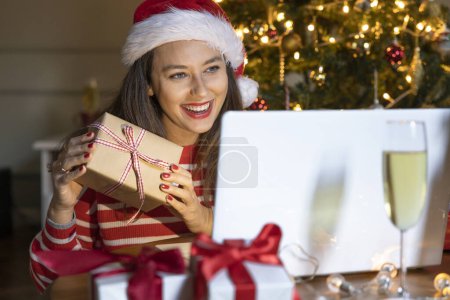 Photo for Cheerful woman holding a gift box while using laptop and making video call on Christmas evenin - Royalty Free Image