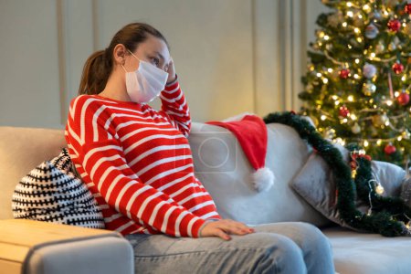 Photo for Young woman home alone on Christmas with Christmas hat and protective face mask. Christmas under the effect of pandemic - Royalty Free Image