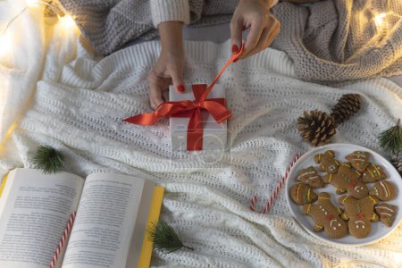 Foto de Las manos de una joven abriendo sus regalos de Navidad sentada en su cama. Celebración de Año Nuevo o cumpleaños - Imagen libre de derechos