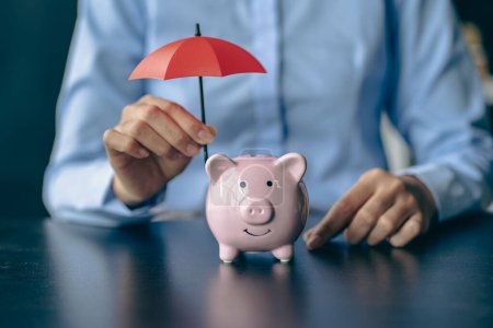 Sparschwein und Frau mit rotem Regenschirm Schützen Sie Vermögenswerte und sparen Sie Geld bei den Käufern der Krankenversicherung. Frau mit kleinem Regenschirm über Sparschwein vor beigem Hintergrund, Nahaufnahme