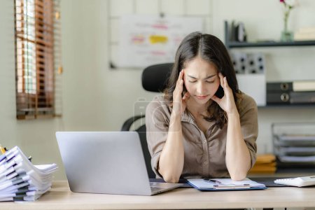 Foto de Asiática mujer de negocios dolor de cabeza durante el trabajo análisis de gráficos financieros con portátil en su escritorio mujer estresada Dolor de cabeza durante las horas extras en la oficina en casa - Imagen libre de derechos