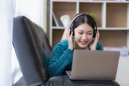 Eine junge Asiatin hört zu Hause auf dem Sofa im Wohnzimmer Musik. Glückliche Frau nutzt Laptop für Videoanrufe mit Freunden im Urlaub