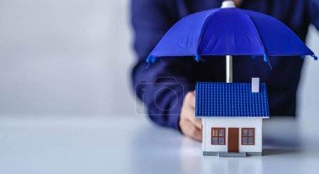 Hand holding a blue umbrella over a mockup house, real estate, insurance and property to protect home and family from harm, insurance concept.