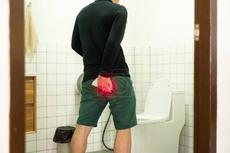 Foto de Joven asiático que sufre de hemorroides, diarrea en el inodoro, cubriendo sus nalgas con puntos rojos en frente del inodoro - Imagen libre de derechos