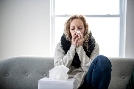 Ill upset young woman sitting on sofa covered with blanket freezing blowing running nose got fever caught cold sneezing in tissue, sick girl having influenza symptoms coughing at home, flu concept