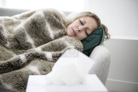 Ill upset young woman sitting on sofa covered with blanket freezing blowing running nose got fever caught cold sneezing in tissue, sick girl having influenza symptoms coughing at home, flu concept