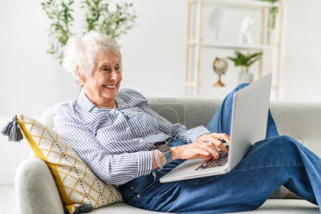Eine lächelnde ältere, graue Seniorin sitzt zu Hause auf dem Sofa