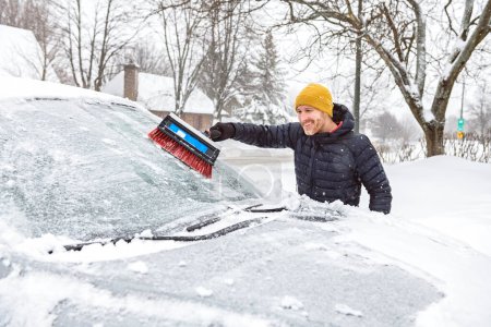 Un jeune homme nettoie sa voiture après une chute de neige par une journée ensoleillée et glacée