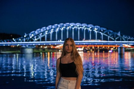 a young woman is strikingly framed with the illuminated Josef Pilsudski Bridge behind her, reflecting over the Vistula River and enhancing the vibrant nightlife of Krakow