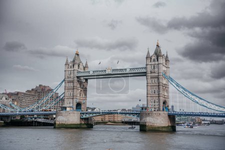 A dramatic view of Londons Tower Bridge under a moody overcast sky, showcasing its distinctive Victorian Gothic architecture and the busy Thames River below