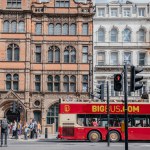 A red open-top sightseeing bus drives past historic buildings with Victorian and Georgian architectural details in London, highlighting the citys rich heritage and tourism