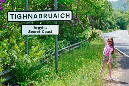 Téléchargez les photos : Jeune fille au panneau Tighnabruaich à l'entrée du village d'Argyll et Bute au Royaume-Uni - en image libre de droit