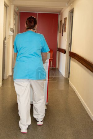 Téléchargez les photos : Infirmière aide femme derrière marche arrière dans le couloir avec chariot - en image libre de droit