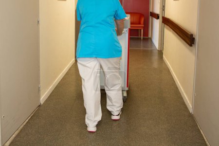 Téléchargez les photos : Infirmière auxiliaire tri blanchisserie dans le couloir de l'hôpital à l'arrière vue marche avec chariot - en image libre de droit