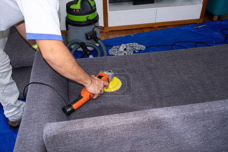 Foto de Hombre con cepillo pulidor eléctrico limpiando un sofá gris. - Imagen libre de derechos