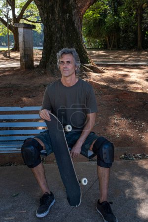 Brazilian skateboarder over 50 years old relaxing on a bench in a square_2.
