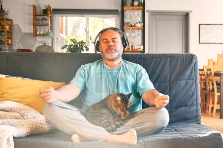 Téléchargez les photos : Mature middle-aged overweight man in wireless headphones relaxing at home with his cat and guided meditation, listening to relaxing music on smartphone and meditating in lotus pose. - en image libre de droit