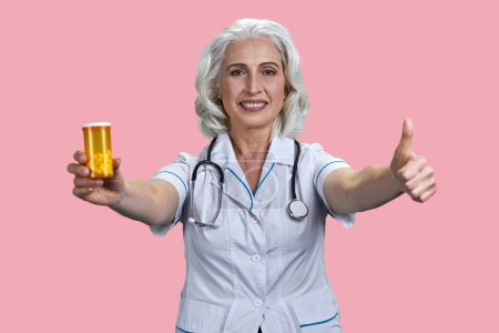 Téléchargez les photos : Portrait de femme âgée heureuse médecin montre bouteille de médicament jaune et pouce vers le haut. Isolé sur fond rose. - en image libre de droit