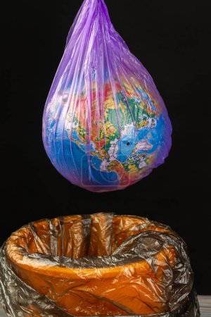 Téléchargez les photos : Globe mondial dans un sac en plastique suspendu au-dessus de la poubelle. Plan vertical isolé sur noir. - en image libre de droit